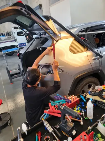 Mechanic repairing a car’s body panel in a workshop, using a tool near the open hatchback.