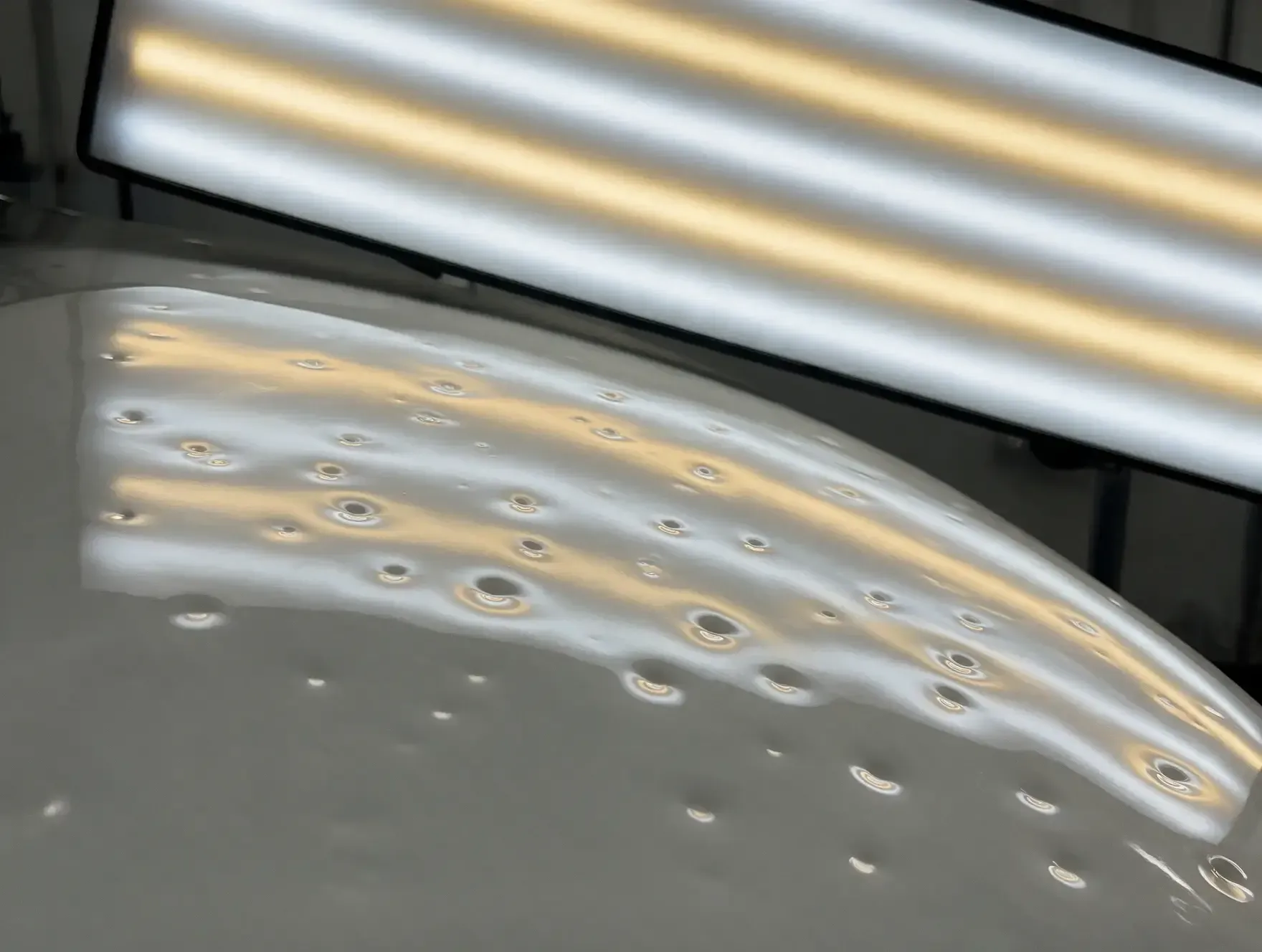 Close-up of a glossy gray curved surface with many small circular dents under bright fluorescent lights.