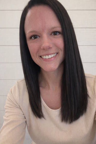 A smiling person with long dark hair wears a beige long-sleeved shirt against a white horizontal paneled background.