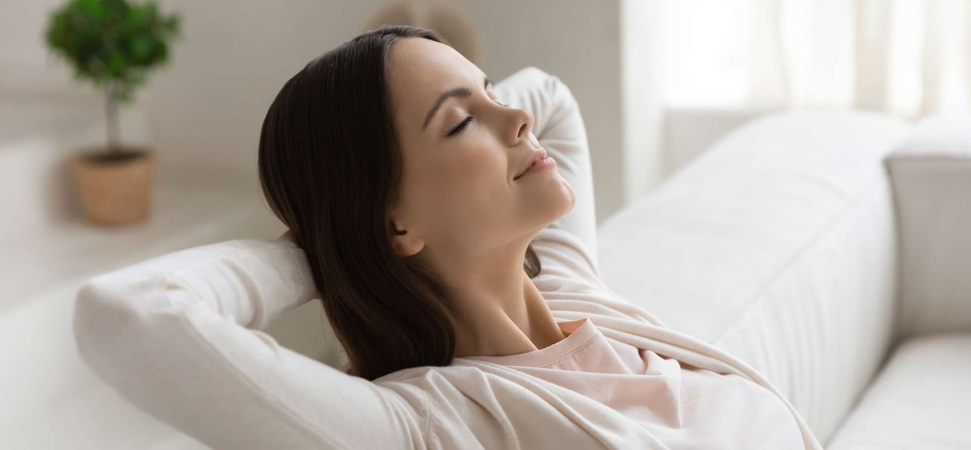 A woman is sitting on a couch with her eyes closed and her hands behind her head representing Breathe Cleaner, Healthier Air at Home