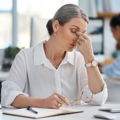 A woman with a cortisol imbalance in a white blouse rubbing her temple at a desk, appearing tired during office work