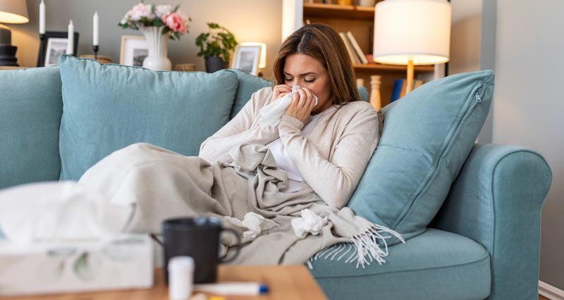 Woman on a blue sofa, wrapped in a blanket, blowing her nose with a tissue, looking unwell.