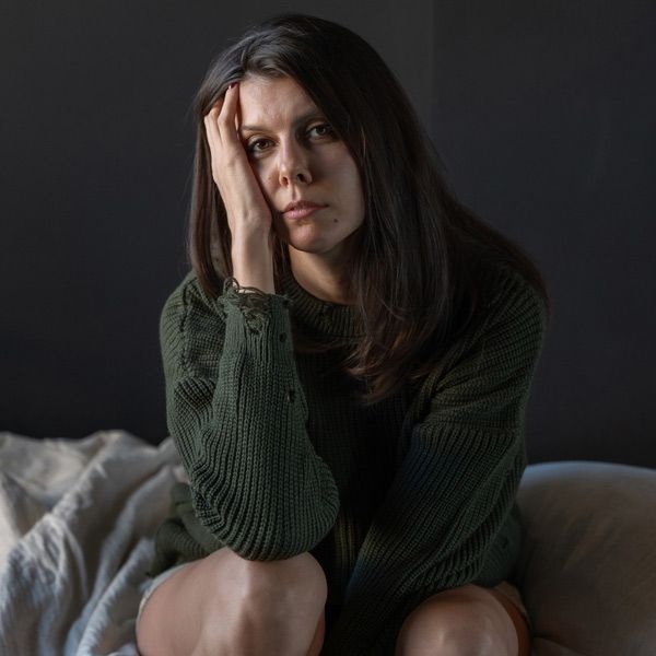 A moody woman with DHEA Imbalance is sitting on a couch, resting her head on her hand with a pensive expression.