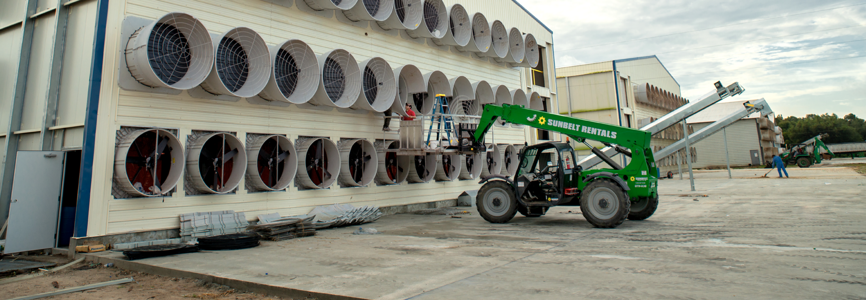 Close-up of an agricultural facility with large ventilation fans on the exterior wall, alongside a forklift and workers performing maintenance
