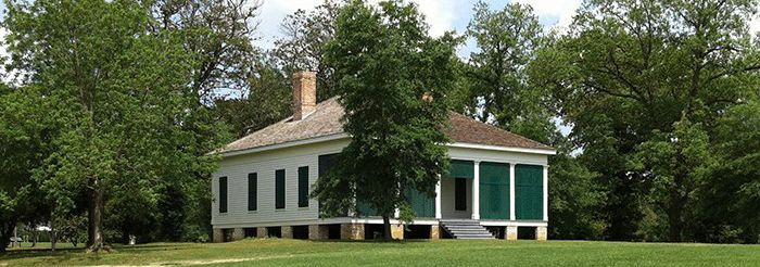 Historic white wooden house with green shutters, elevated on pillars, surrounded by lush trees and an open grassy field.
