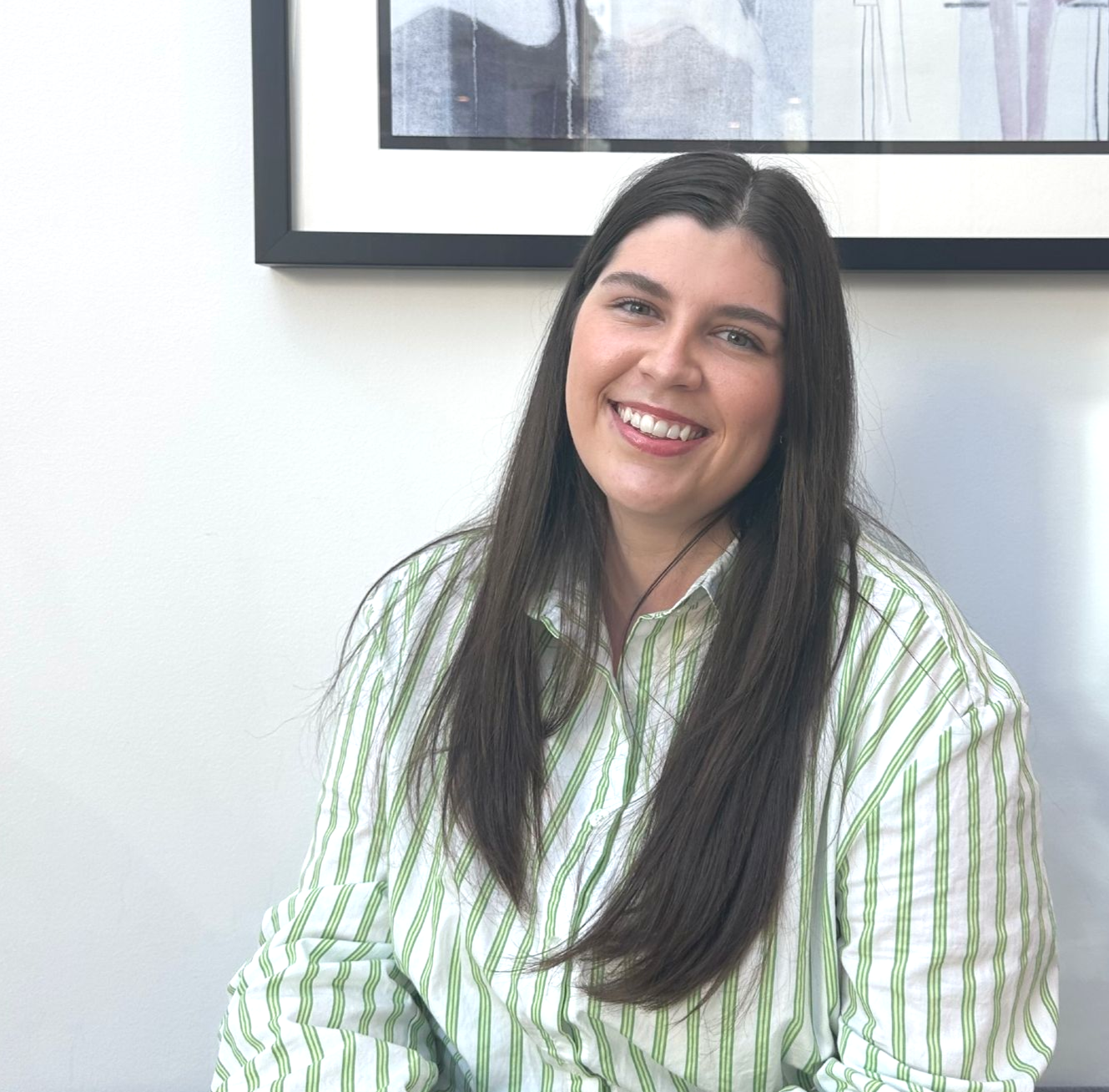 Woman with long dark hair smiles, wearing a green-striped shirt, seated in front of art.