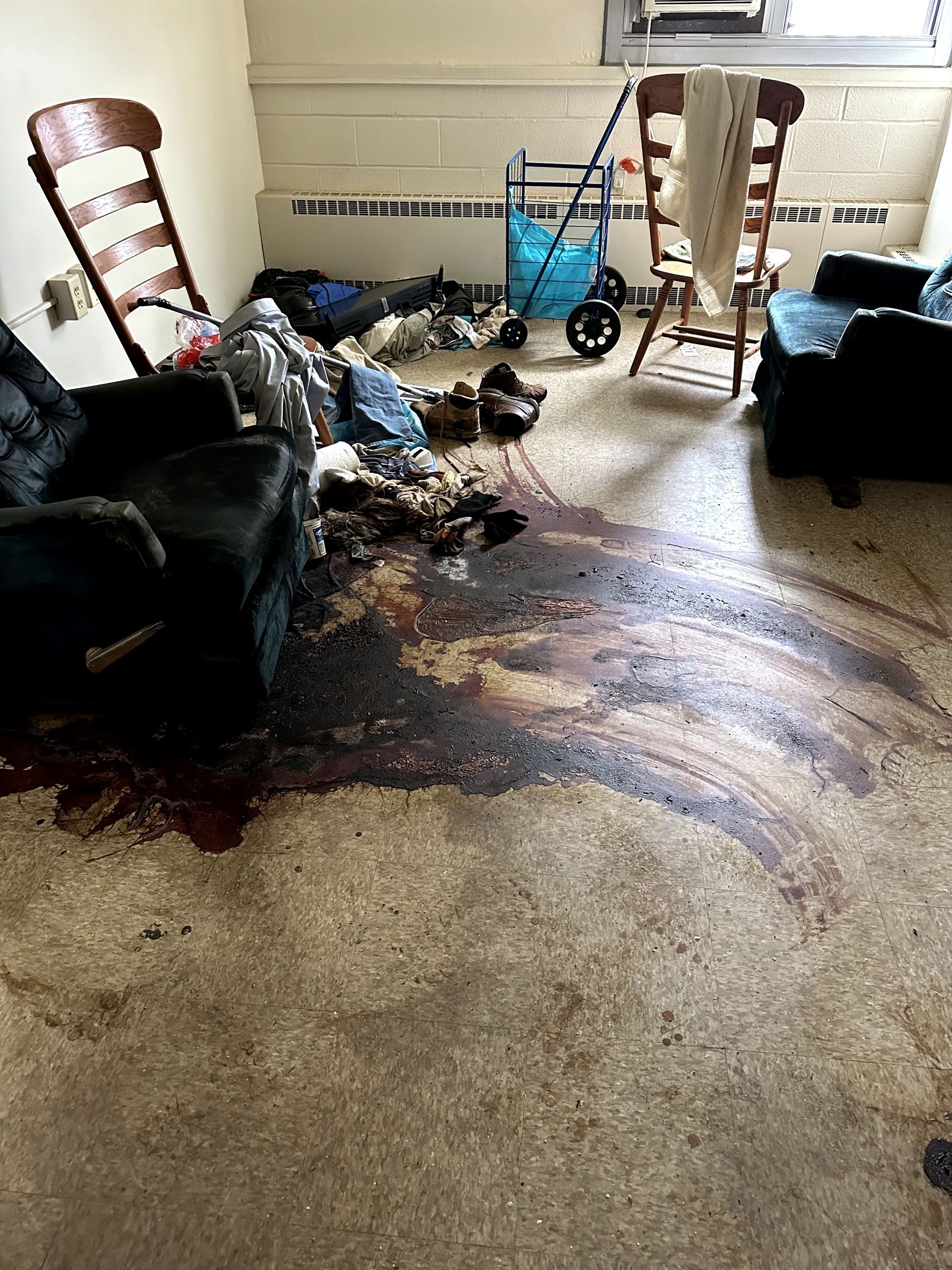 Room with dark stains on floor, debris, damaged furniture. Two chairs, a cart, and a window are visible.