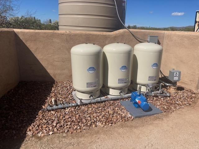 Three beige water tanks and a pump system on gravel in an outdoor setting, with a large water tank in the background.