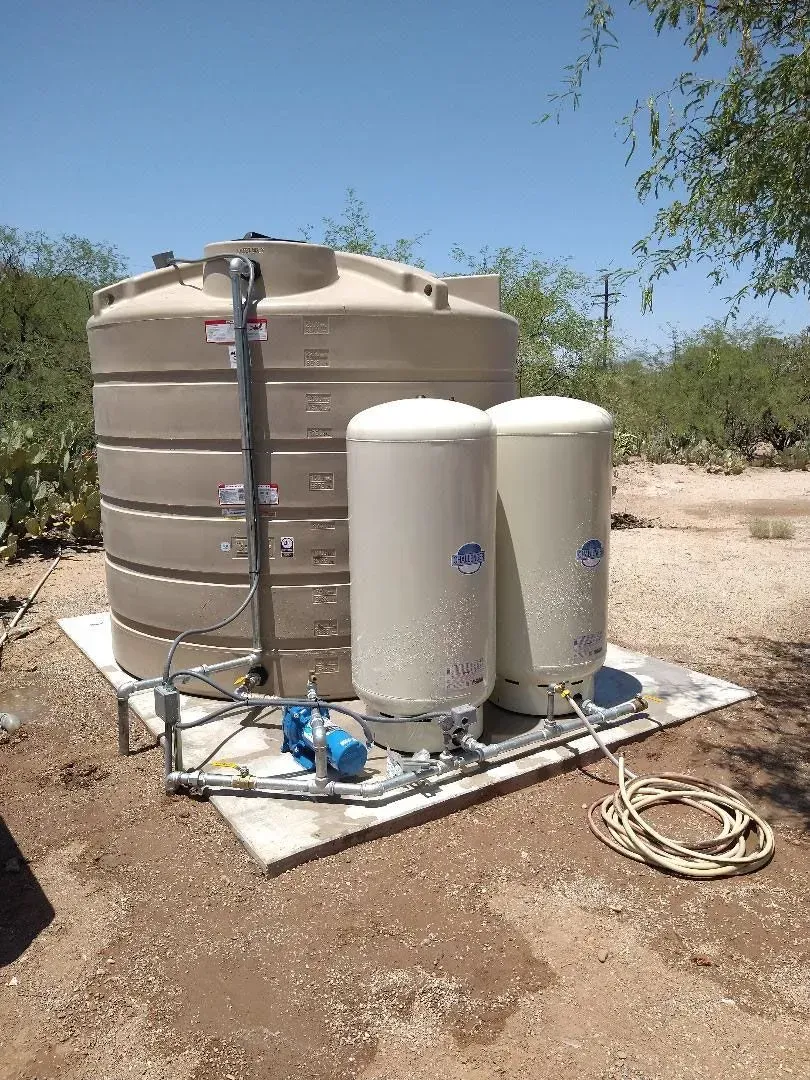 Water storage and filtration system outdoors, beige tanks, blue pump, and piping on concrete pad, sunny setting.