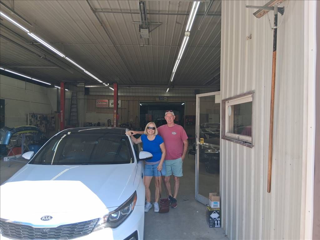 Two people standing beside a white car inside a garage, with a corrugated metal wall on the right