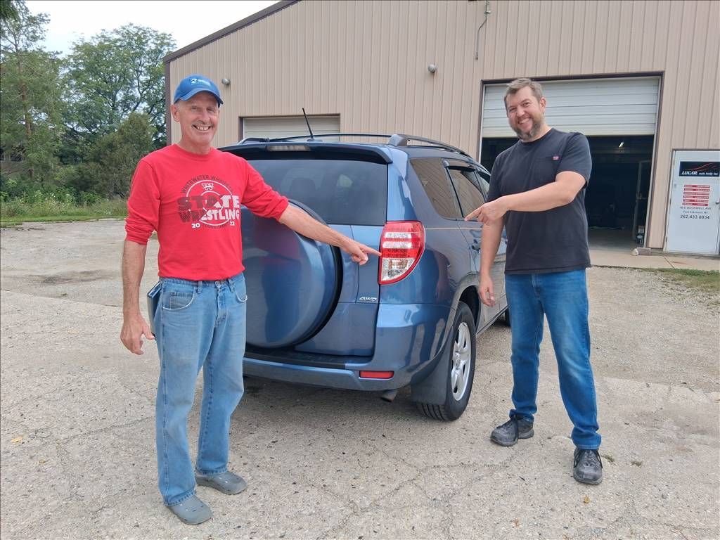 Two men standing beside a blue SUV in front of a garage, one pointing at the rear of the vehicle.