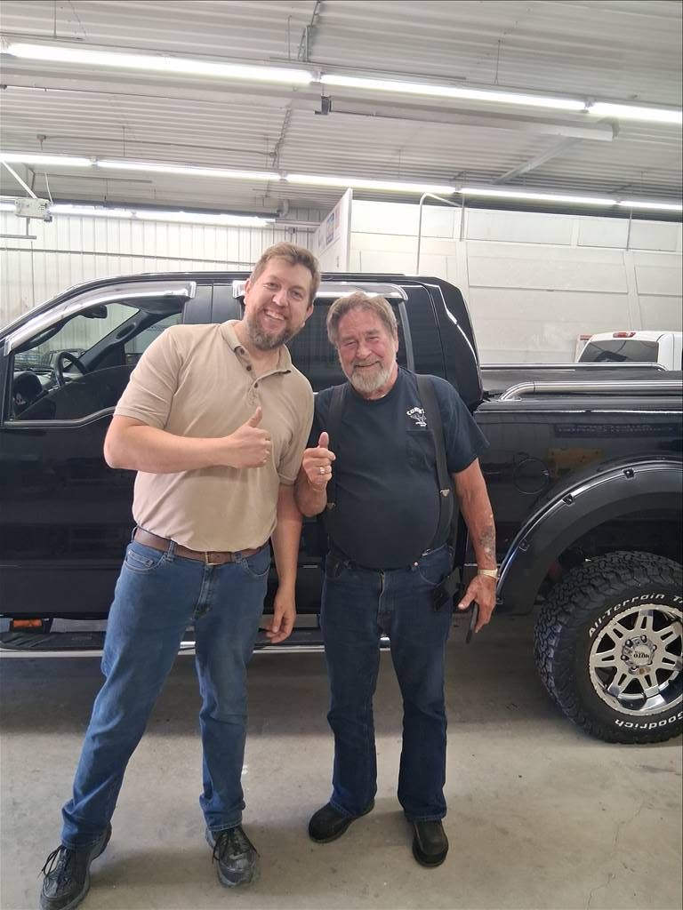 Two men stand beside a black pickup truck in a garage, smiling and giving thumbs up.