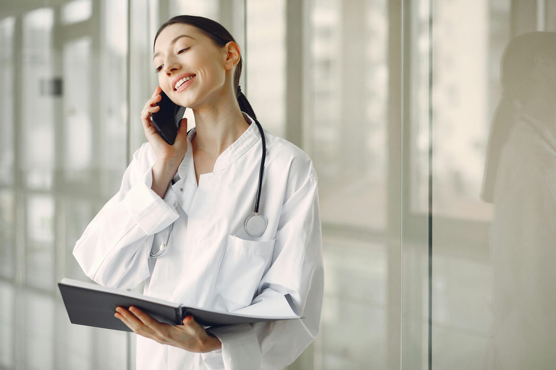 Una doctora con bata blanca sonríe, con un teléfono pegado a la oreja y una libreta. De pie en un pasillo bien iluminado.