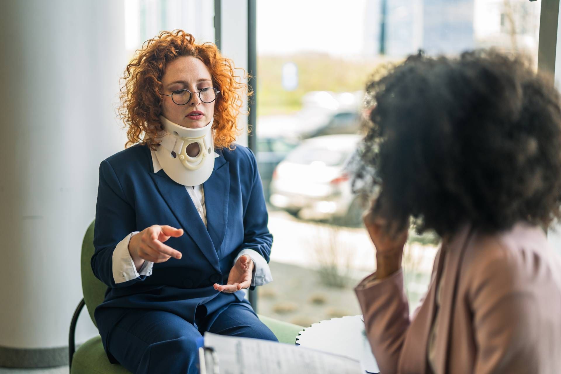 Two people in a clinic, one wearing a neck brace speaking to another seated across from them.