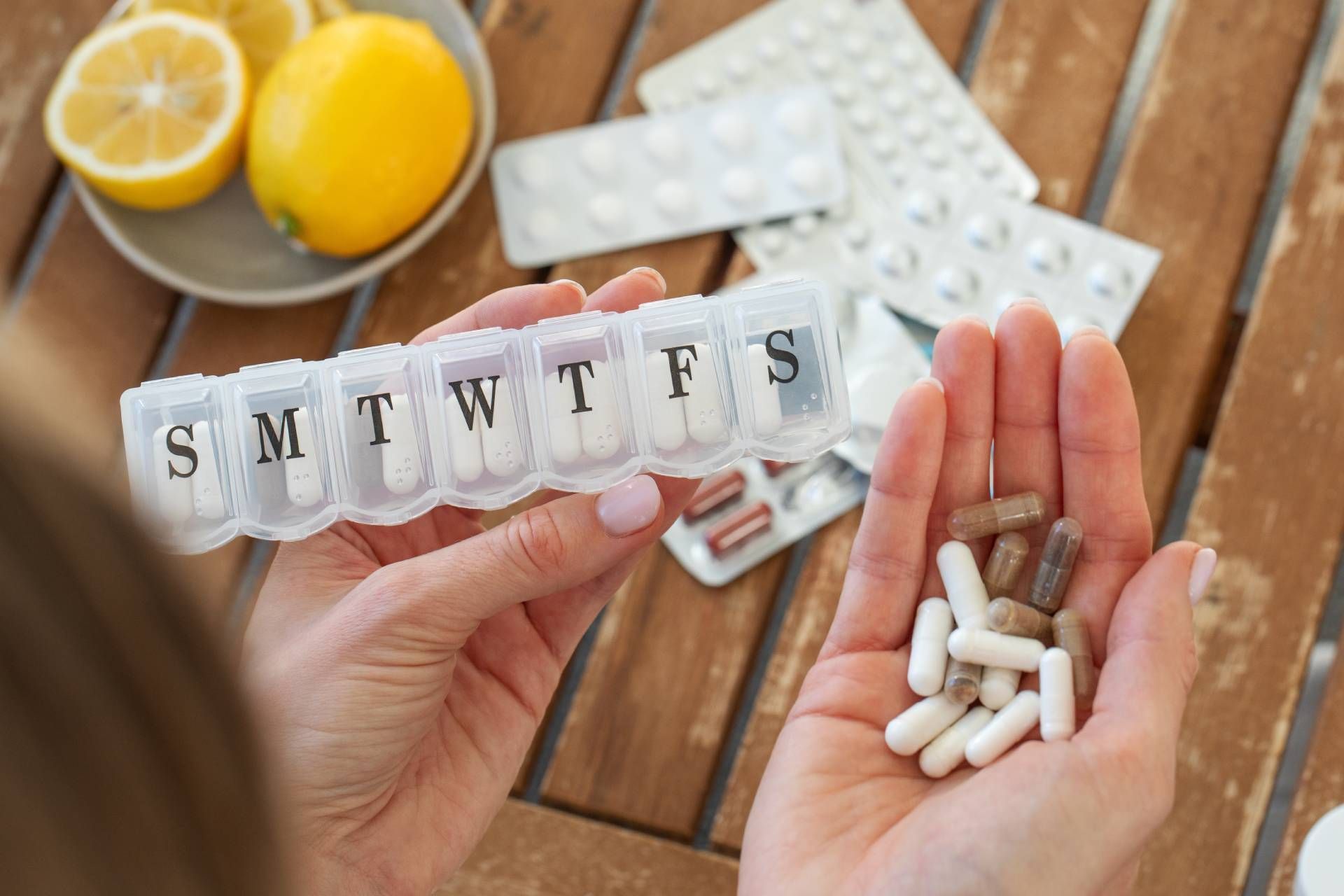 Hands holding a weekly pill organizer and capsules beside a lemon drink on a wooden table