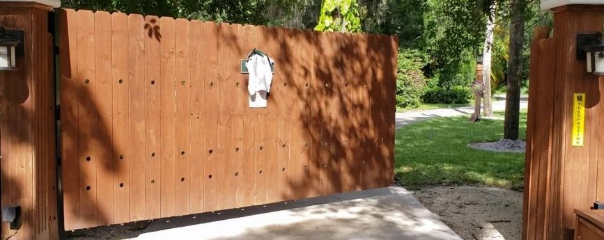 A wooden gate is open to a driveway with trees in the background.