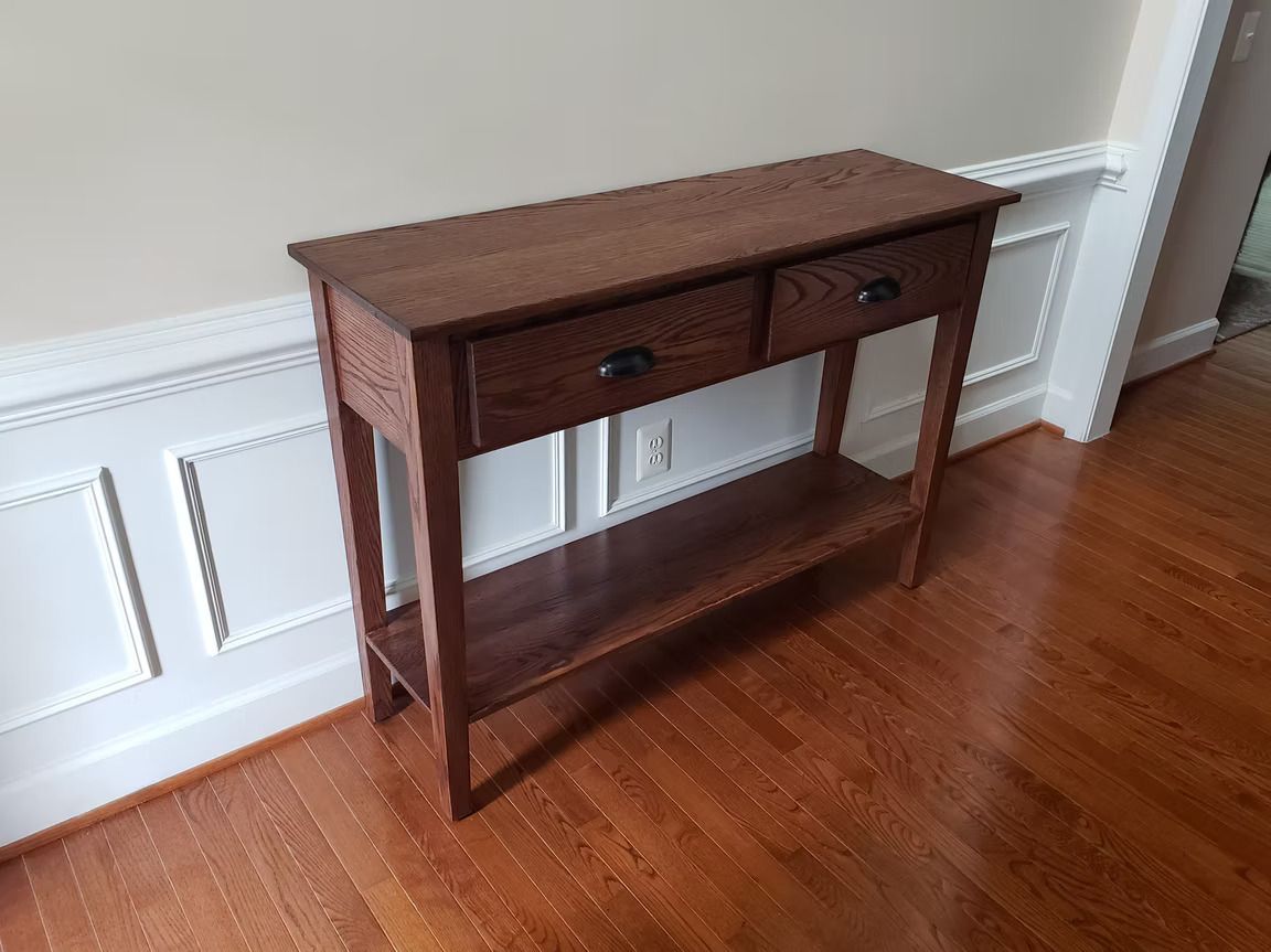 Brown wooden console table with drawers against a white paneled wall on a hardwood floor