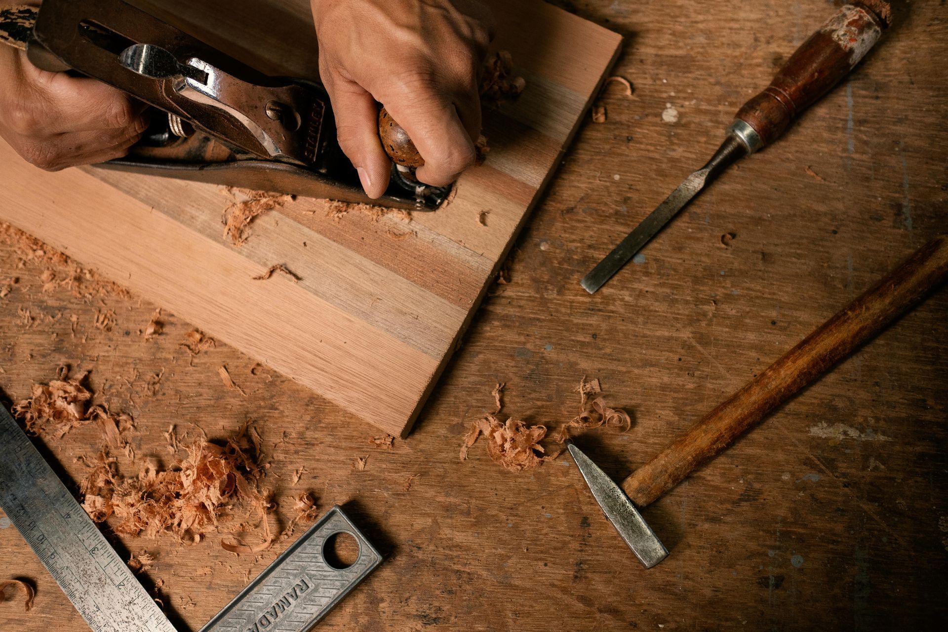 Hands carving wood with chisels and a hammer on a workbench, surrounded by wood shavings