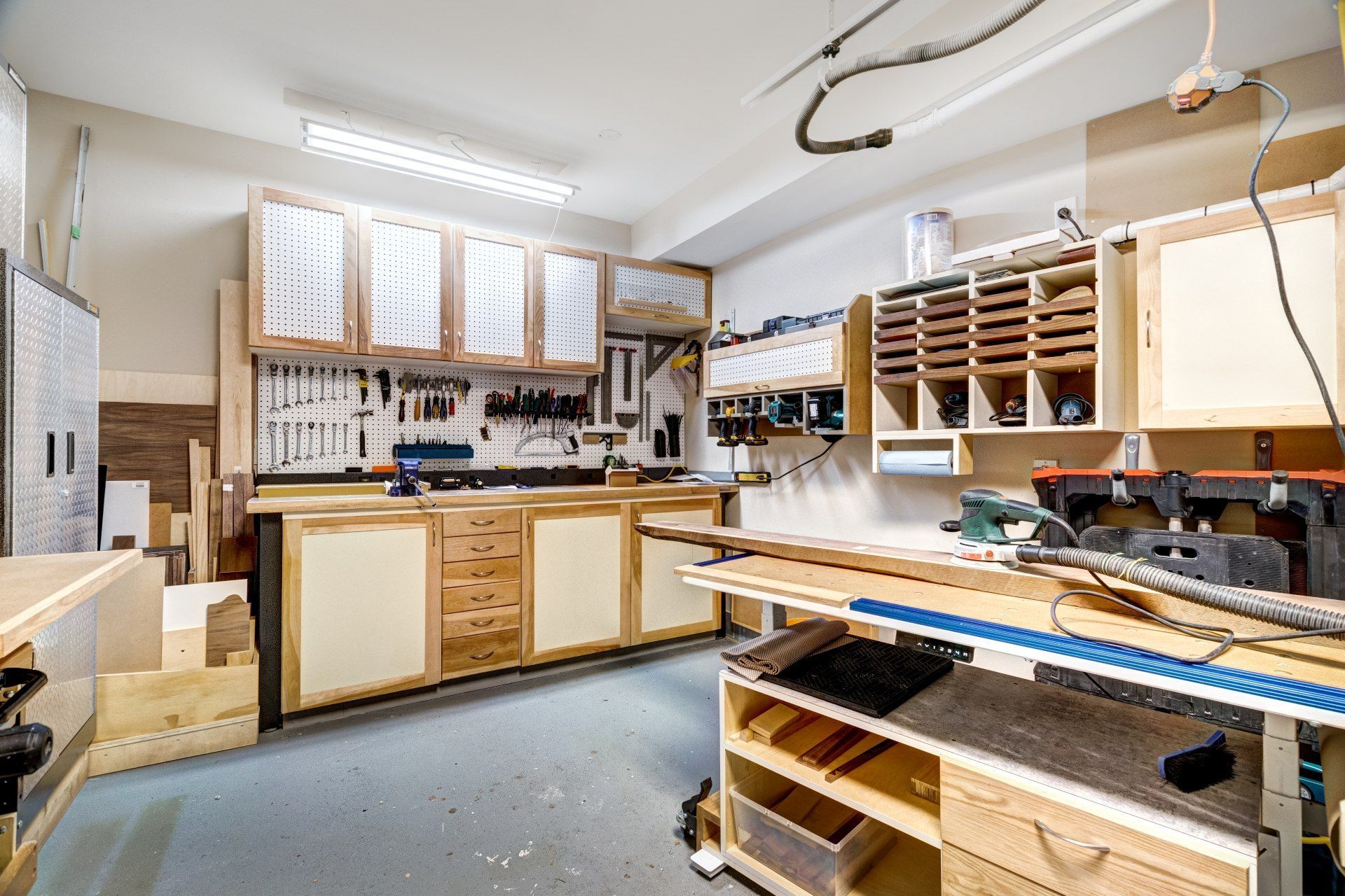 Bright woodworking shop with workbenches, cabinets, tools, and saws under fluorescent lights