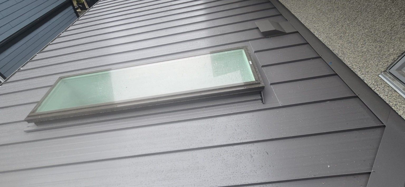 Grey raised seam metal roofing beside a skylight, vent and stucco wall