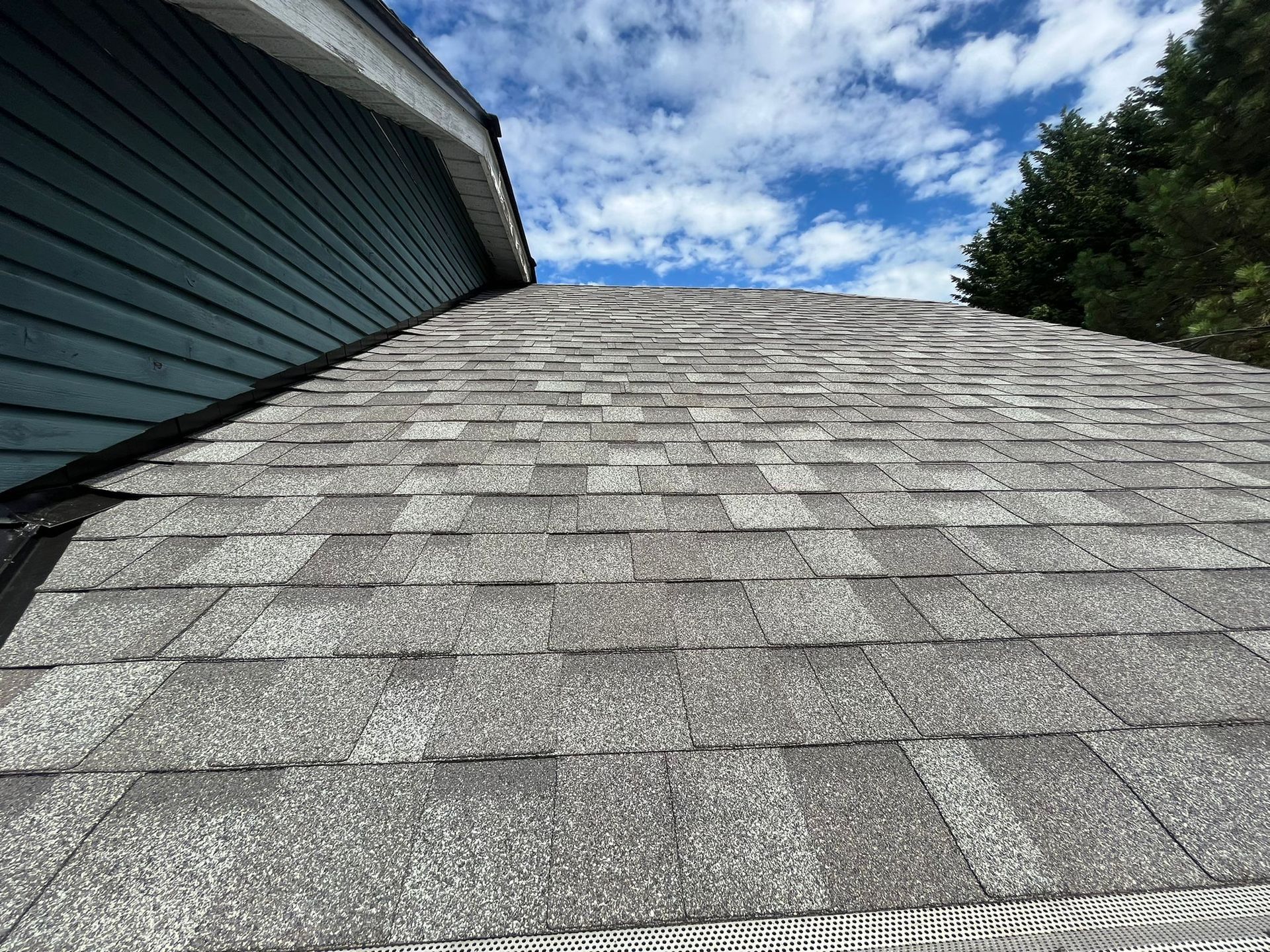A close-up view of a shingled roof next to blue-grey siding under a bright, partly cloudy sky.