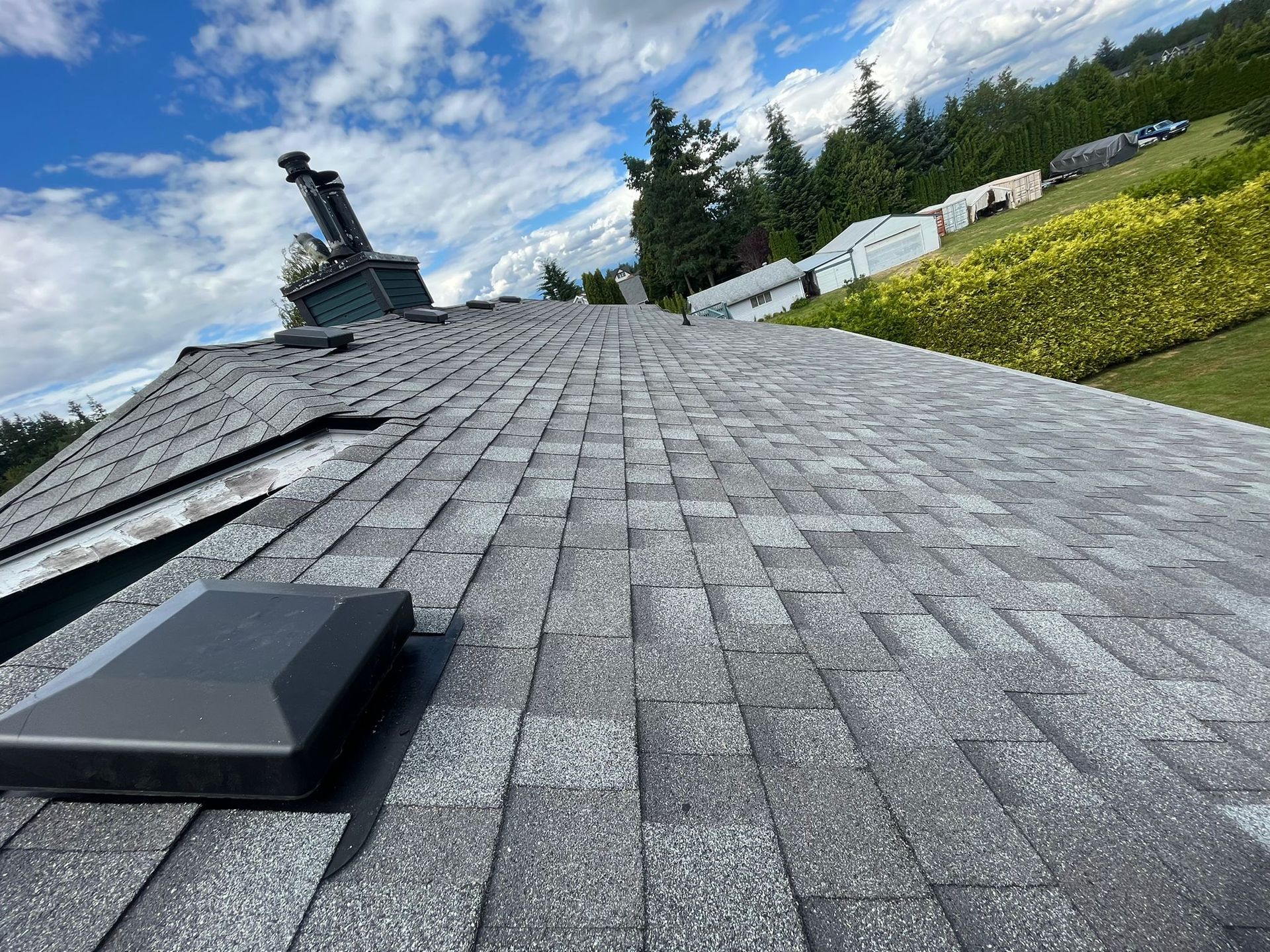 A view looking down the ridge of a gray asphalt shingle roof with a chimney and a black ventilation vent under a blue sky.