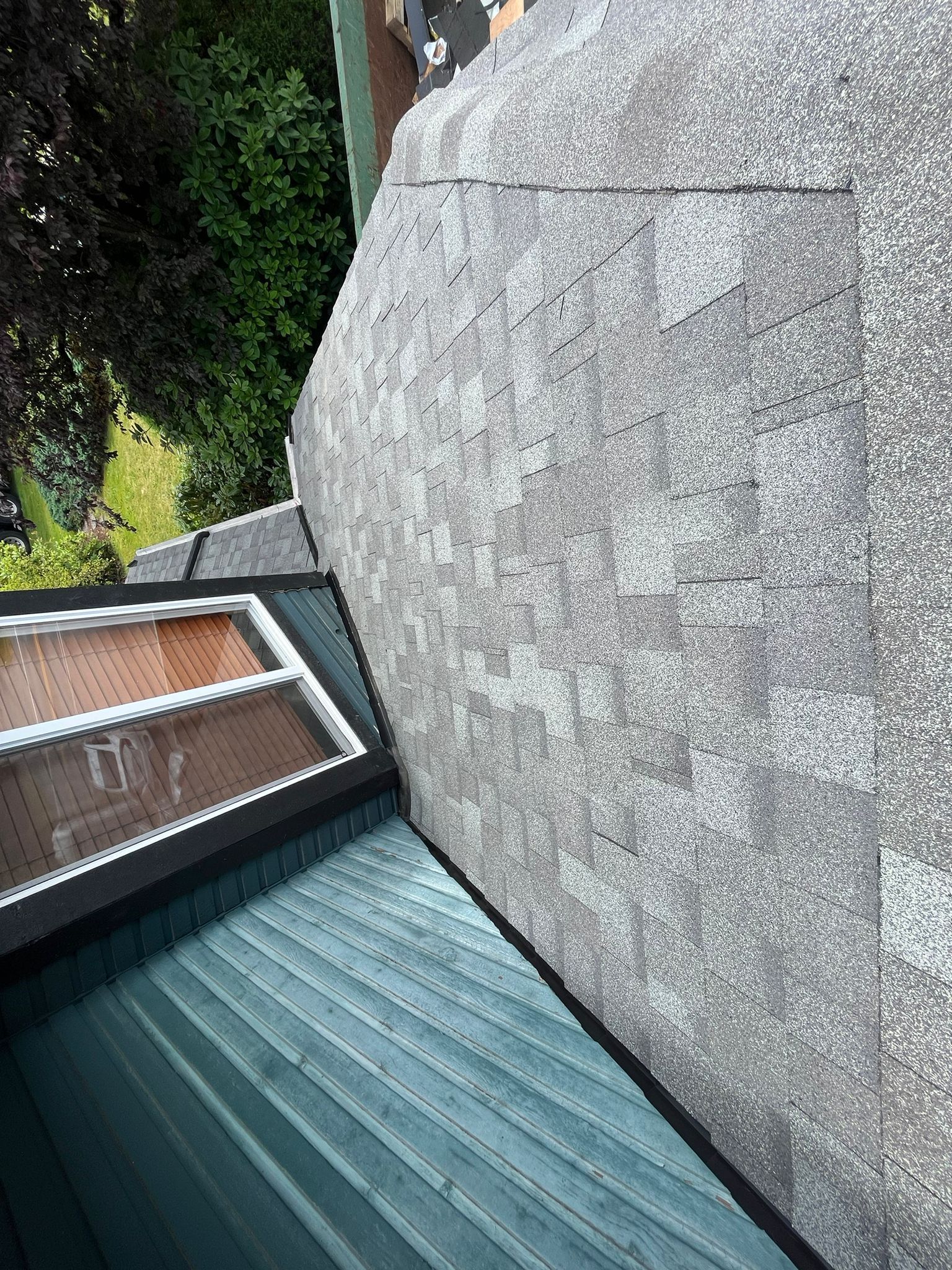 High-angle view of a roof with grey shingles meeting a green corrugated metal roof and a small window structure.