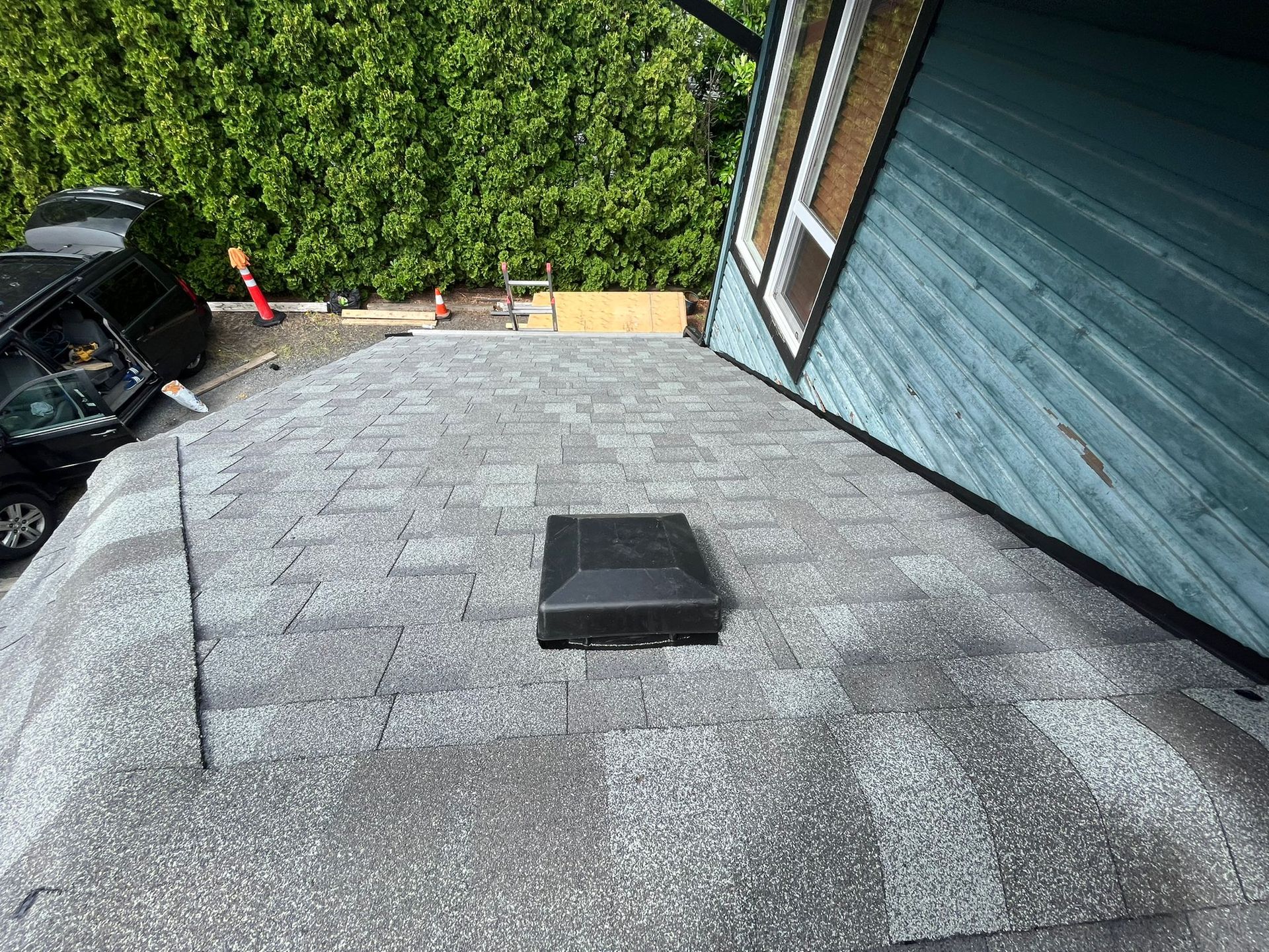 A high-angle view of a gray asphalt shingle roof with a black square roof vent, next to a blue-sided house wall.