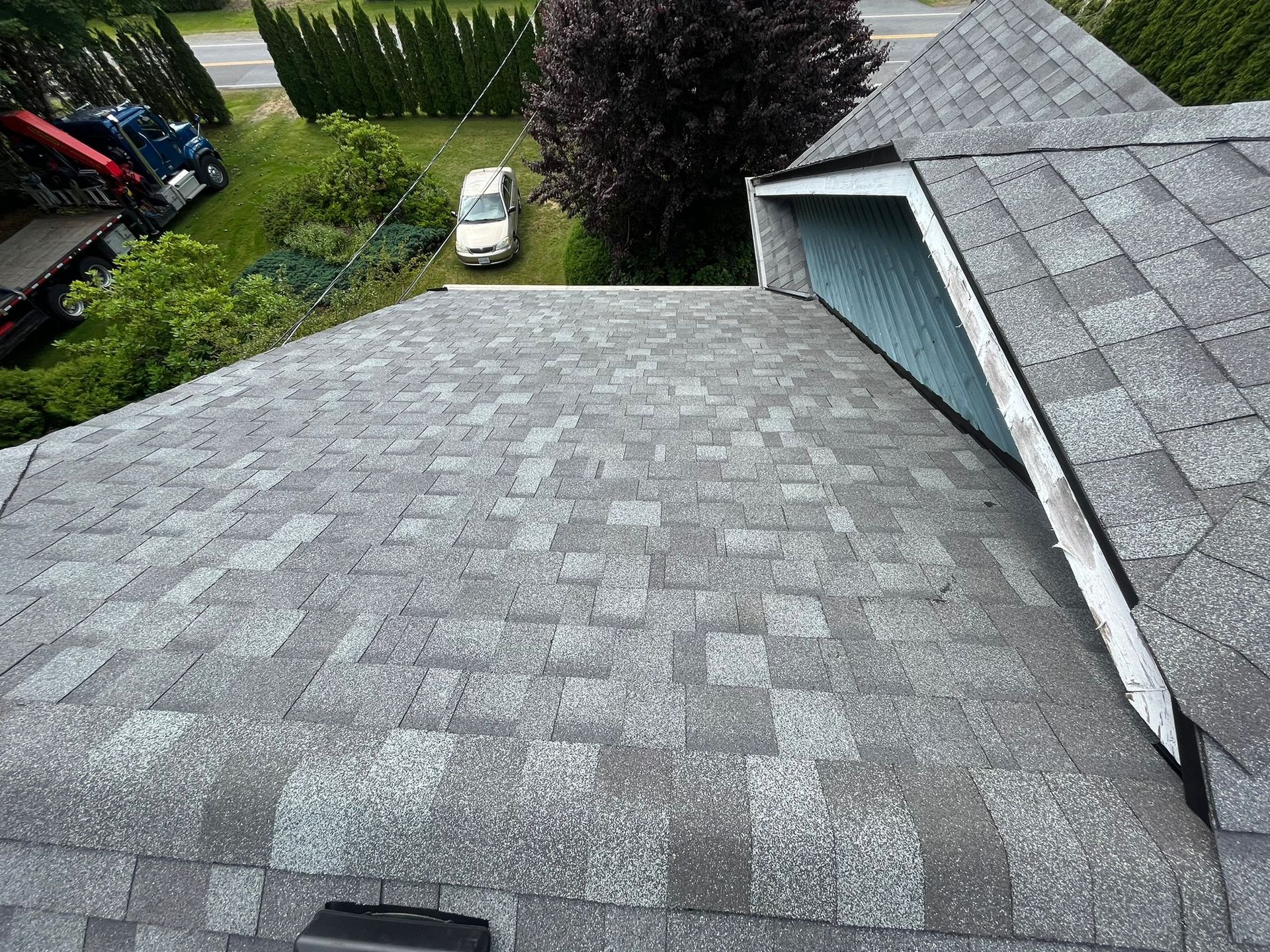A high-angle view of a gray shingled roof with a dormer window, overlooking a grassy yard with a car and trees.