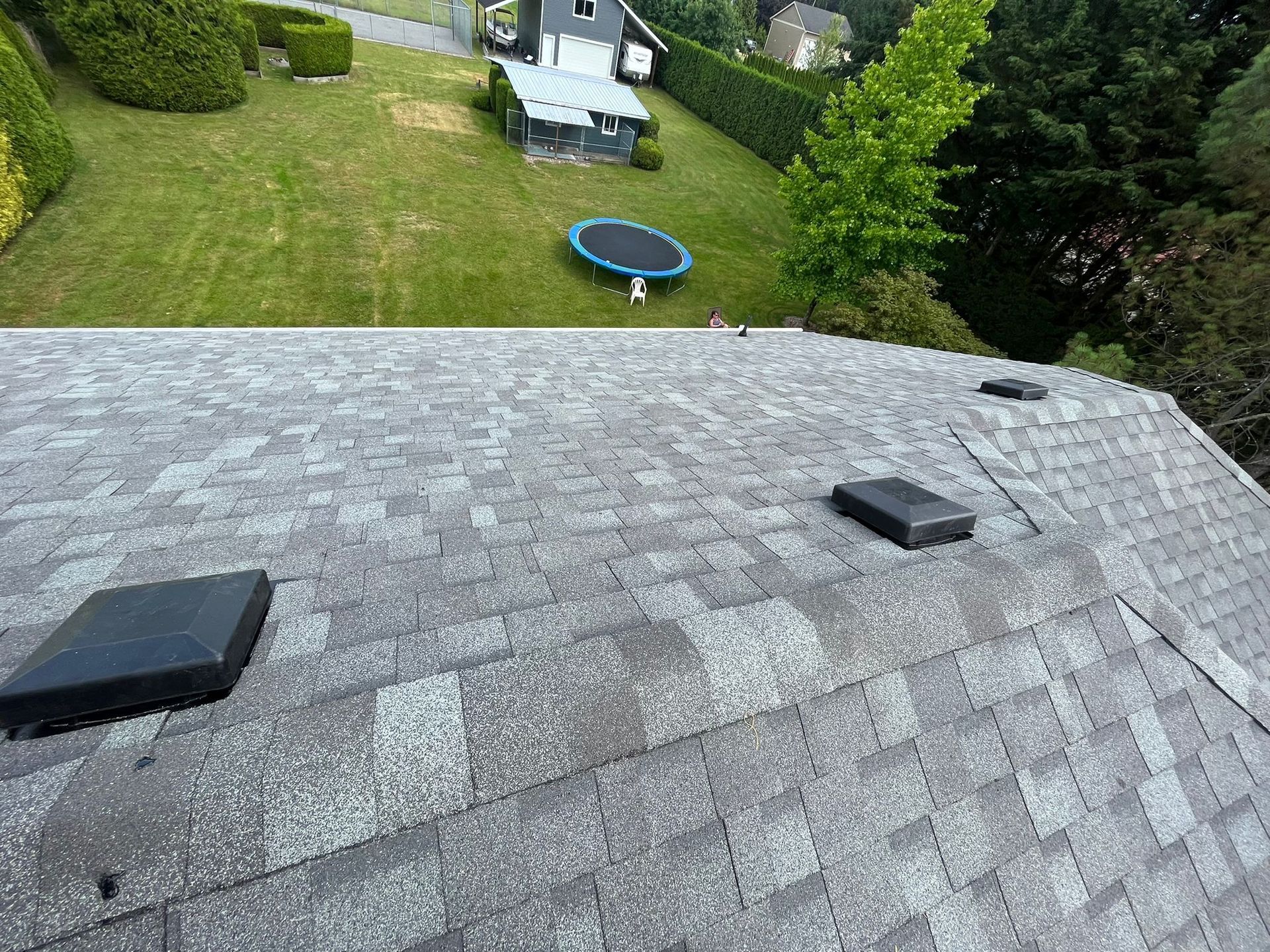 An elevated view of a gray asphalt shingle roof with three black ventilation vents, overlooking a yard with a trampoline.