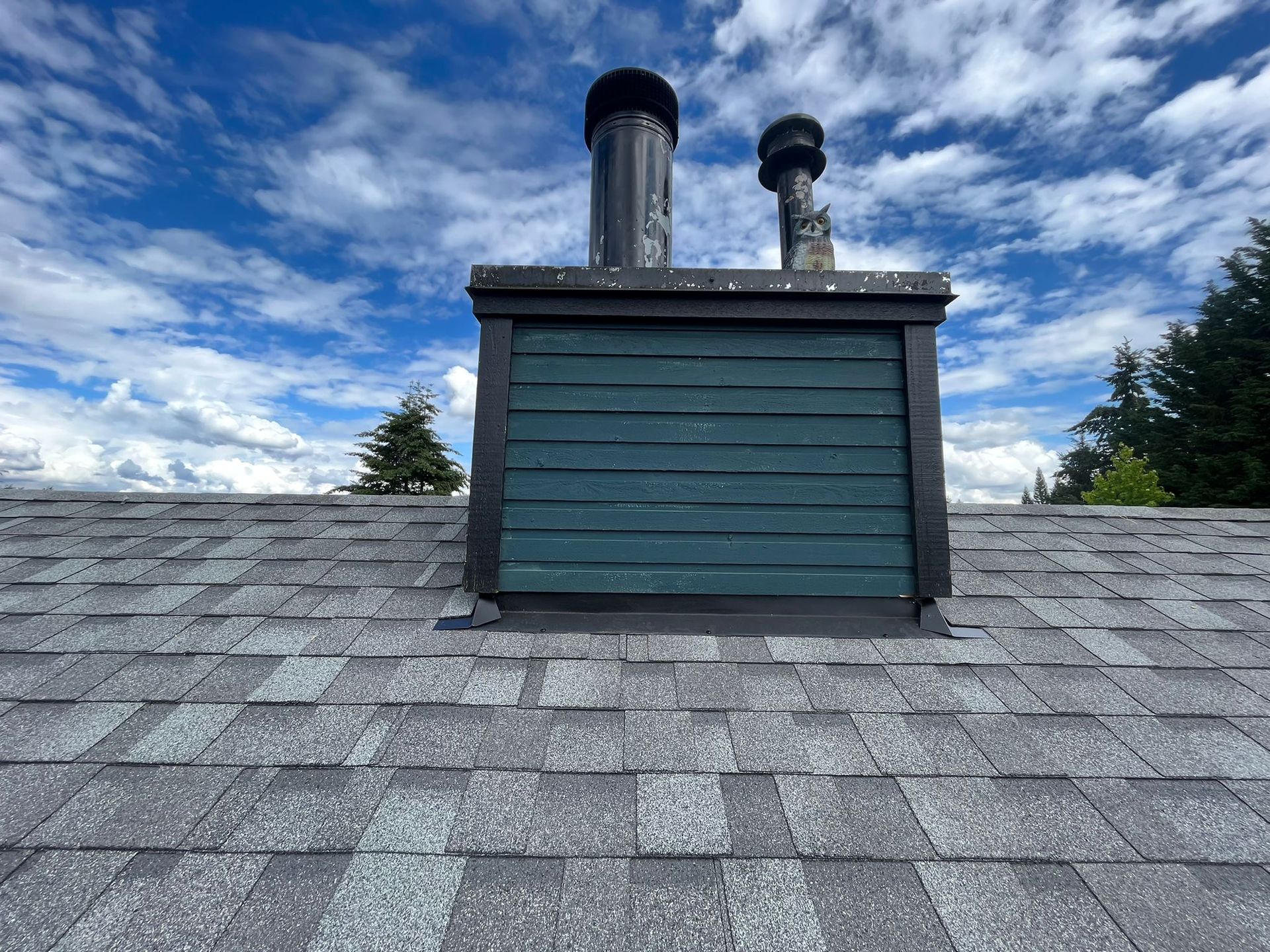 A chimney with green siding and two black metal caps stands on a gray shingled roof under a blue, cloudy sky.