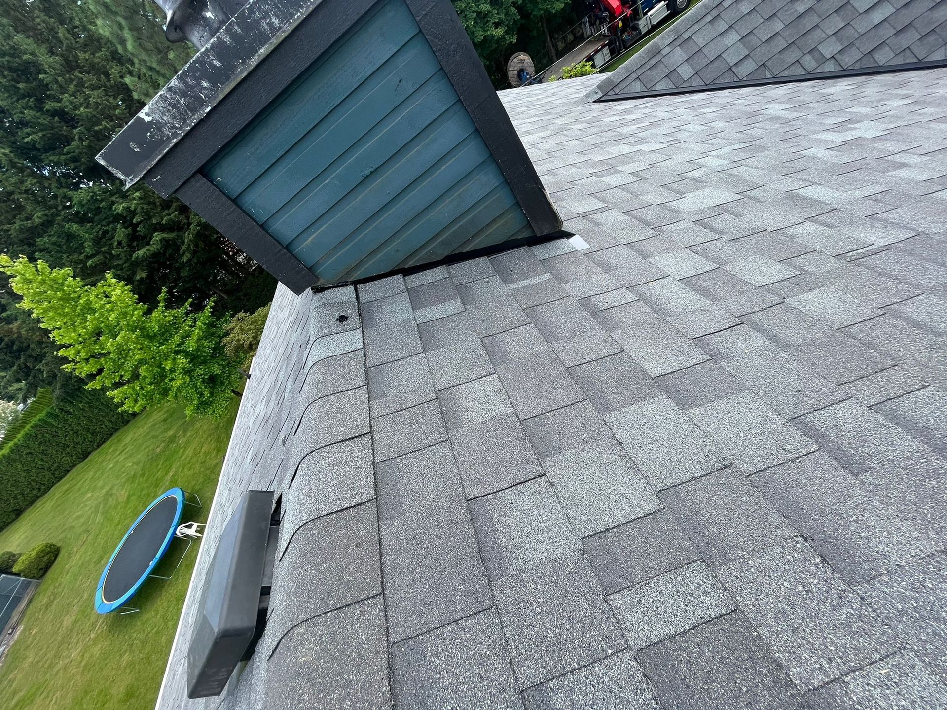 A high-angle view of a gray asphalt shingled roof with a dark-trimmed chimney, a vent, and a green yard with a trampoline.