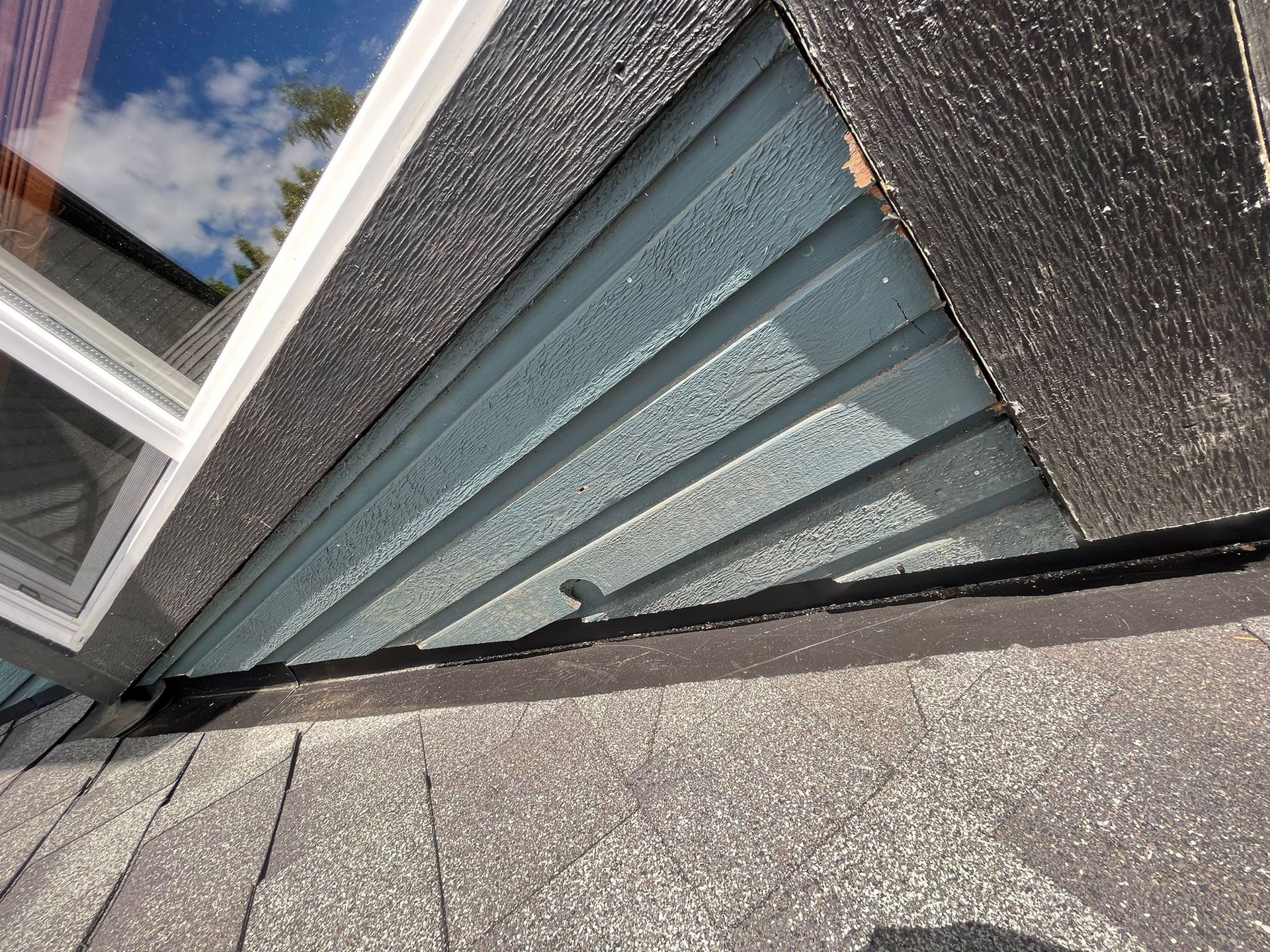 A close-up view of a shingled roof meeting a wall with horizontal blue siding and dark, textured trim.