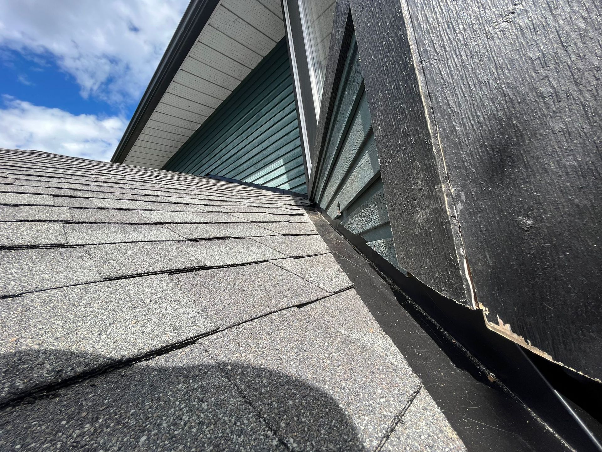 A close-up view of a roof valley where grey shingles meet a black metal flashing against a dark exterior wall.