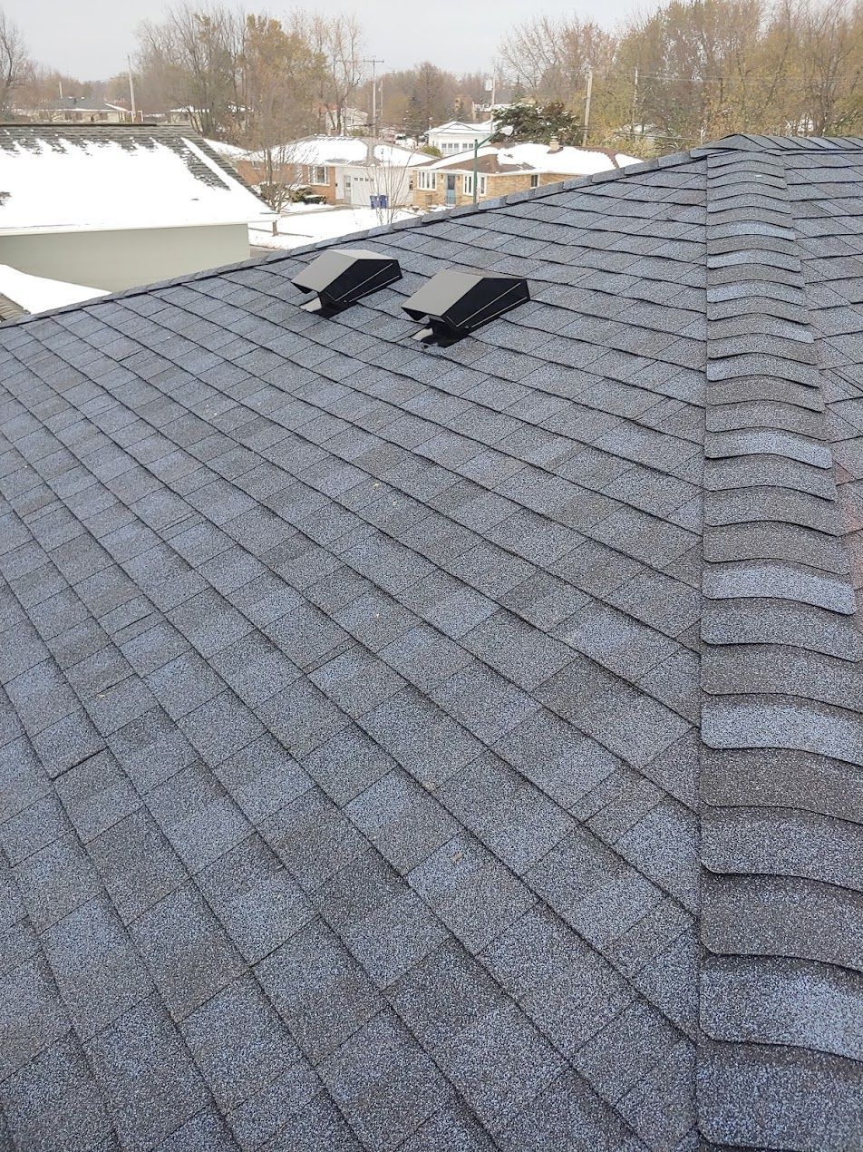 A gray shingled roof covered in light snow features two black static roof vents installed on a sloped section.