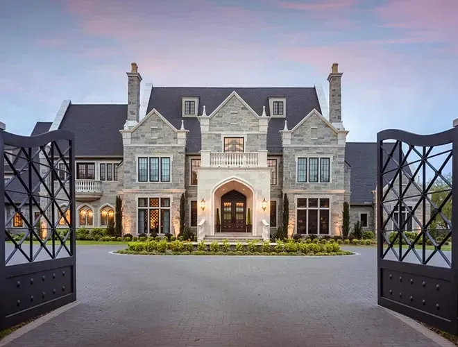 Stone mansion with open black gates, long driveway, and dusk sky.