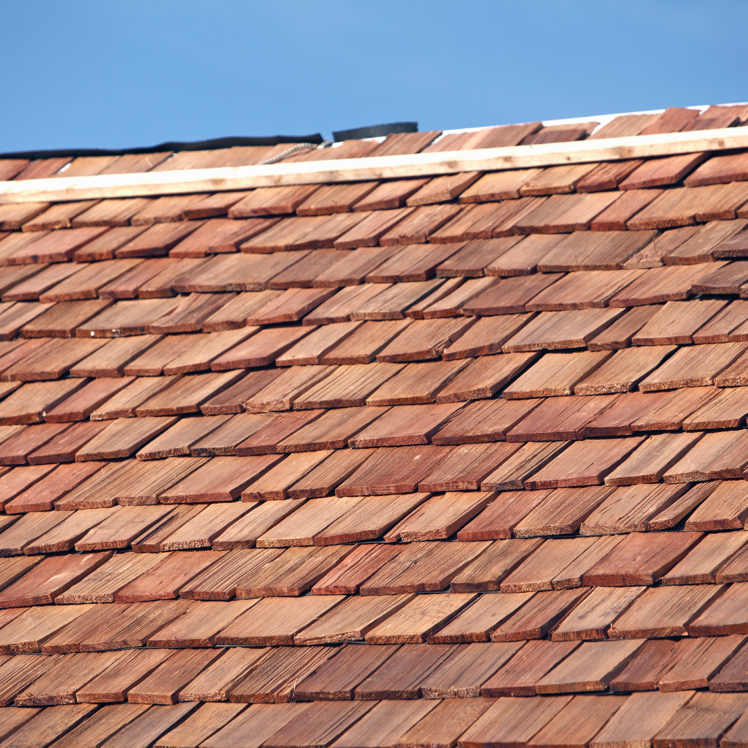 A close-up view of a roof covered in brown wooden shingles, featuring a horizontal wooden ridge cap against a blue sky.
