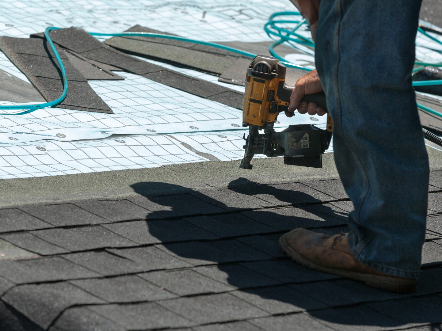 A worker uses a nail gun to install dark asphalt shingles on a roof over a light blue underlayment.