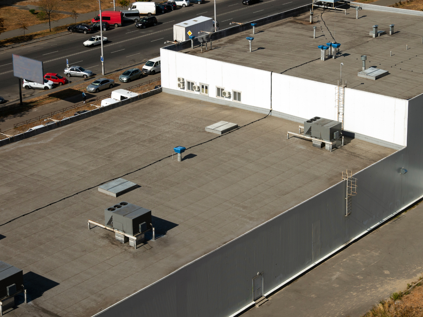 An aerial view of a flat, gray commercial building roof with several HVAC units, next to a road with moving cars.