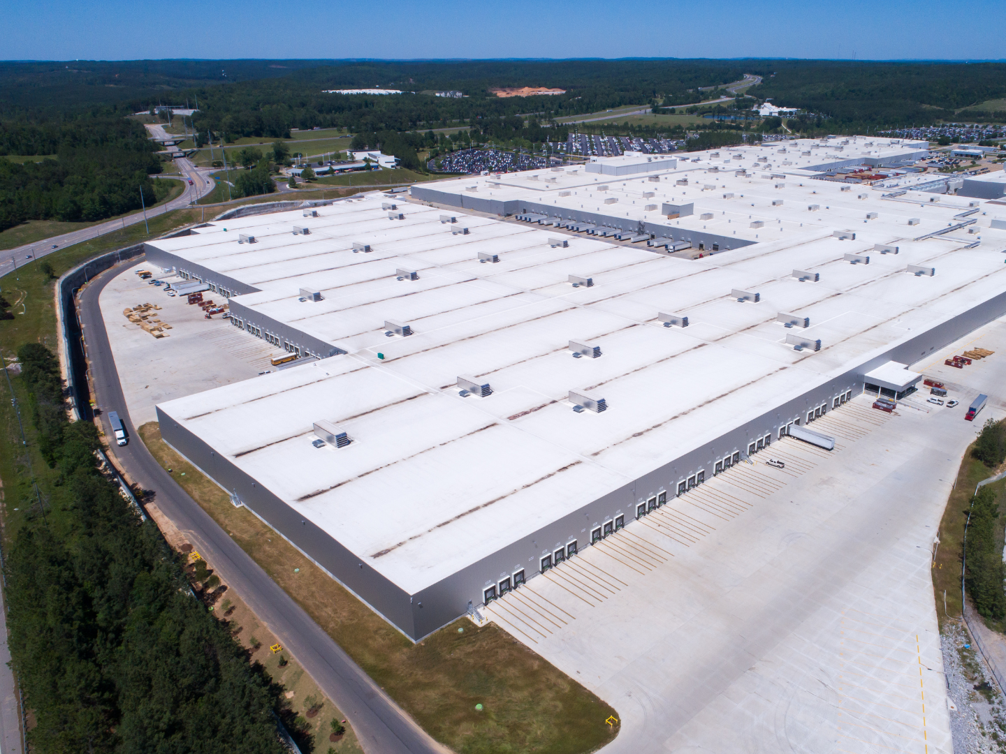 Aerial view of a large, flat-roofed industrial warehouse building surrounded by a parking lot and forest.