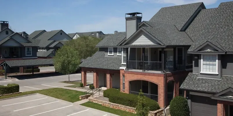 Multi-story apartment buildings with brick and dark siding exteriors, grey shingled roofs, and a parking lot in front.