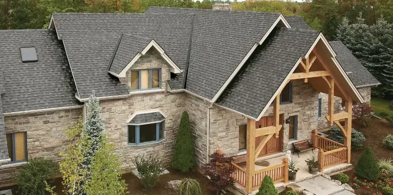 A stone house with a complex dark grey shingled roof, a timber-framed front porch, and surrounding evergreen landscaping.