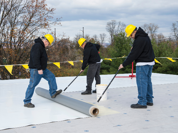 Three construction workers in yellow hard hats and black hoodies install roofing material on a flat roof.