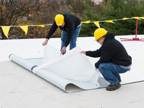 Two workers in yellow hard hats roll out white roofing underlayment on a flat roof.