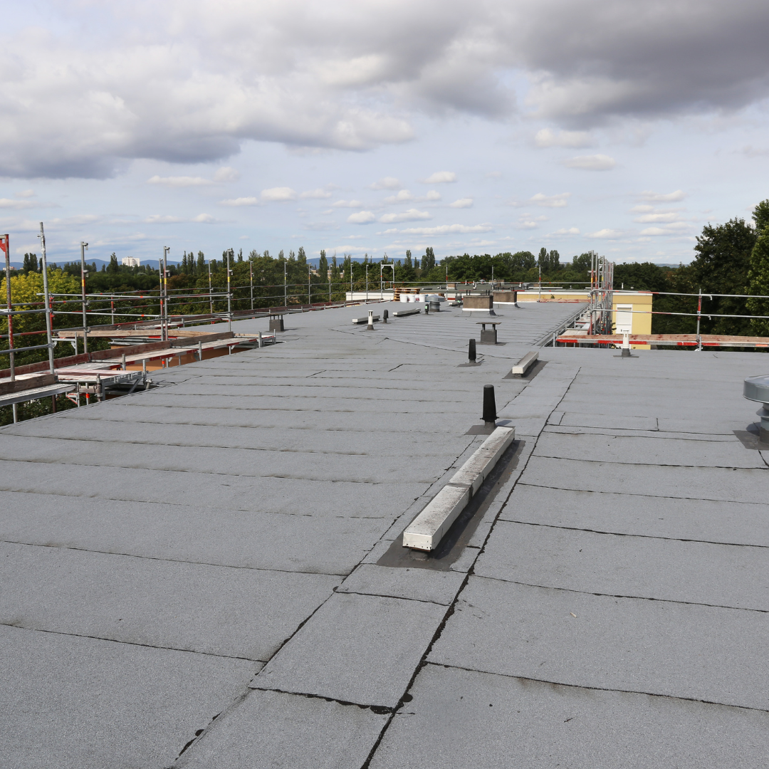 A flat rooftop covered in grey material, featuring a few small vents and surrounded by metal construction scaffolding.