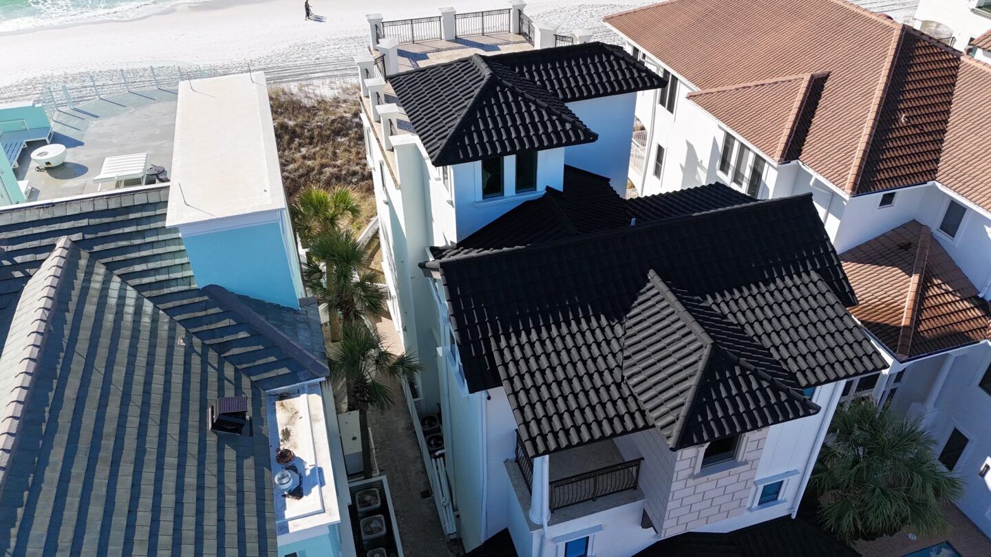 Aerial view of a coastal beach house with a black roof next to a house with a brown roof near the sandy shore.
