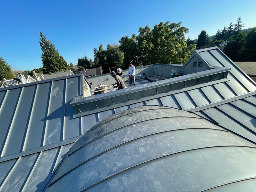 Two workers on a standing seam metal roof, with one positioned on a curved roof section and another on a flat platform.