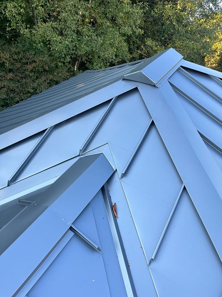 A close-up of a blue metal roof with raised seams and a hip section, set against a background of green trees.