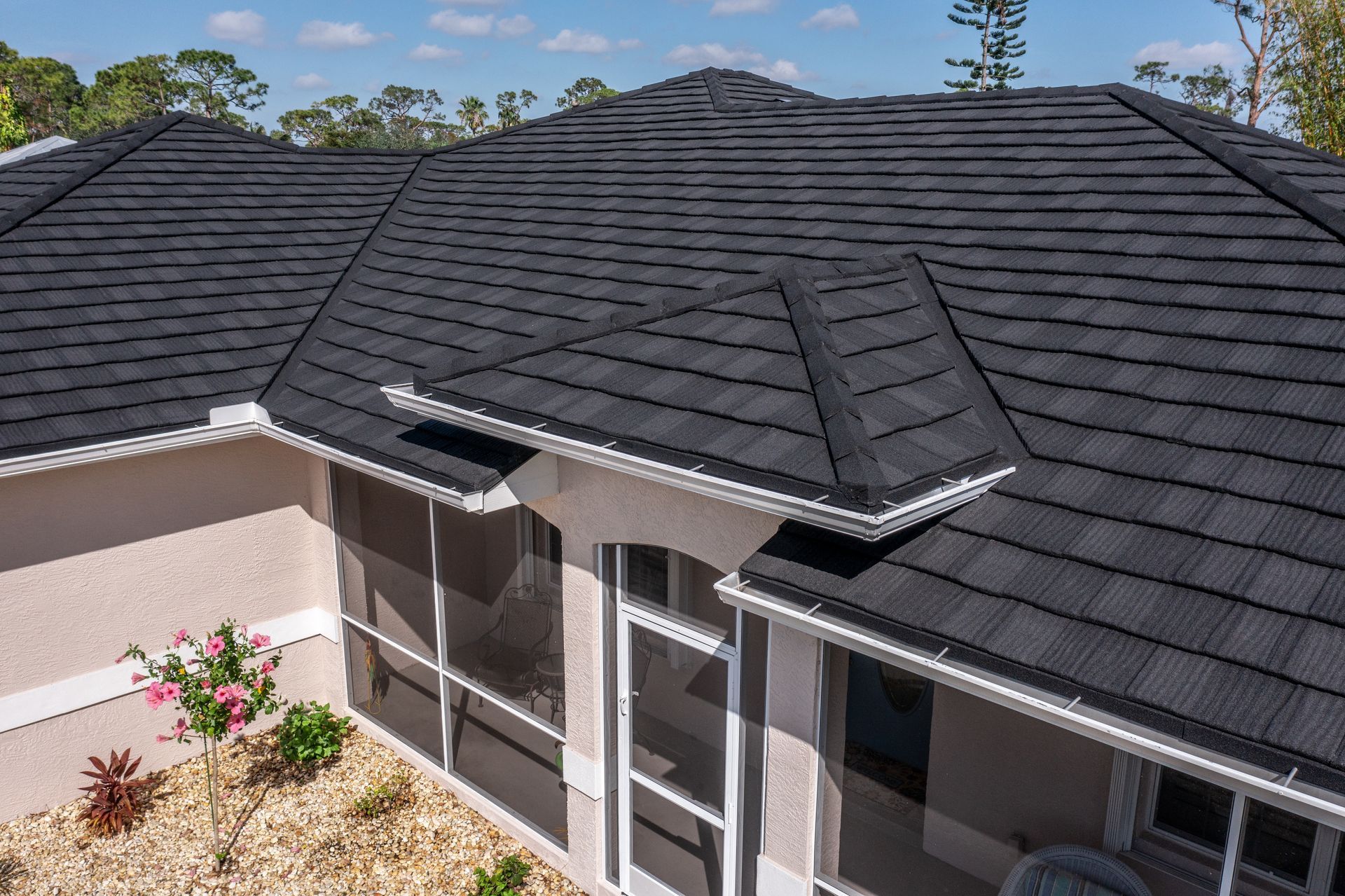 A high-angle view of a dark shingled roof on a beige house with a screened-in porch area and landscaping below.