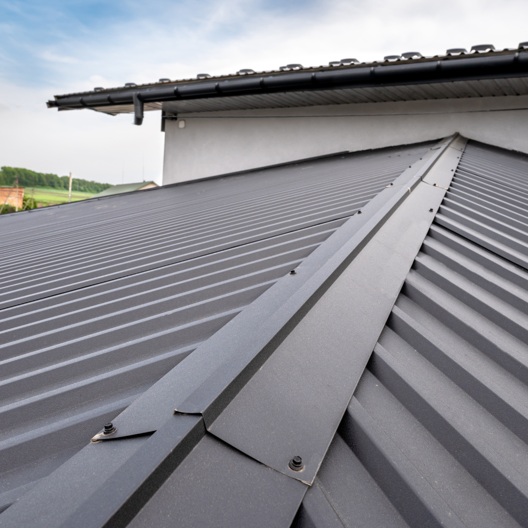 Close-up of a gray metal roof with ridges and a black ridge cap. Black gutters and white siding visible.